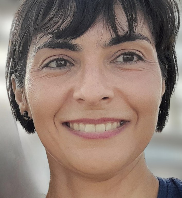 Professora Ana Carolina Soliva Soria. Mulher com cabelos castanho-escuros e franja penteada para o lado. Tem olhos castanhos e usa óculos redondos na cor marrom. Está com camisa vermelha. No fundo, parede de tijolos aparentes com parte de poster com imagem de um moinho, nas cores azul e rosa.
