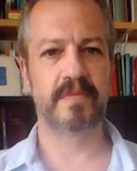 Professor Fernão de Oliveira Salles dos Santos Cruz. Homem com cabelos grisalhos curtos. Barba grisalha cobrindo a parte inferior do rosto. Está com camisa branca. No fundo, estante com livros.