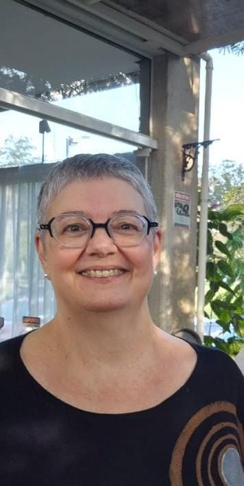 Professora Marisa da Silva Lopes. Mulher com cabelos avermelhados, lisos e curtos. Tem olhos castanhos, utiliza óculos retangulares na cor dourada. Está com blusa rosa e sorriso alegre. 